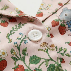 Puede incluir: Primer plano de una tela rosa con un estampado de fresas, hojas y flores blancas. Un bot&oacute;n blanco est&aacute; centrado en la tela. Un rat&oacute;n de dibujos animados con peto es visible cerca del cuello.