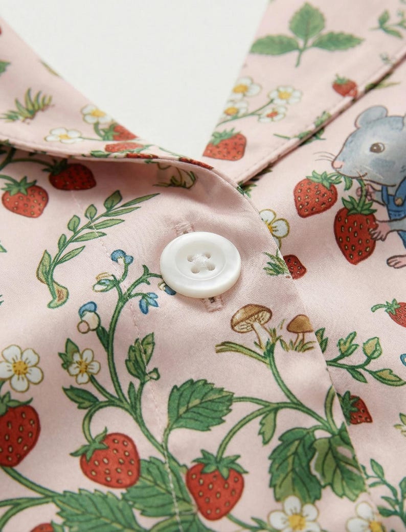 Puede incluir: Primer plano de una tela rosa con un estampado de fresas, hojas y flores blancas. Un bot&oacute;n blanco est&aacute; centrado en la tela. Un rat&oacute;n de dibujos animados con peto es visible cerca del cuello.