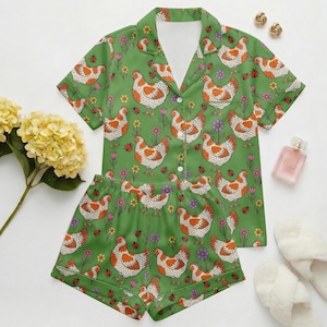 Puede incluir: Conjunto de pijama verde con un estampado repetido de gallinas blancas y naranjas, flores y mariquitas. La parte superior de manga corta y los pantalones cortos están hechos de un material suave. El conjunto incluye una camisa con cuello abotonada y pantalones cortos a juego.