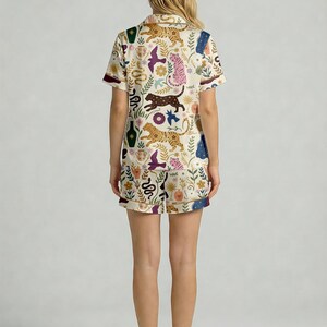 Pode incluir: Conjunto de pijama creme com um padr&atilde;o repetido de tigres, p&aacute;ssaros, cobras e elementos florais. A parte superior de manga curta e os cal&ccedil;&otilde;es t&ecirc;m uma borda contrastante. O design inclui cores castanho, rosa e azul.