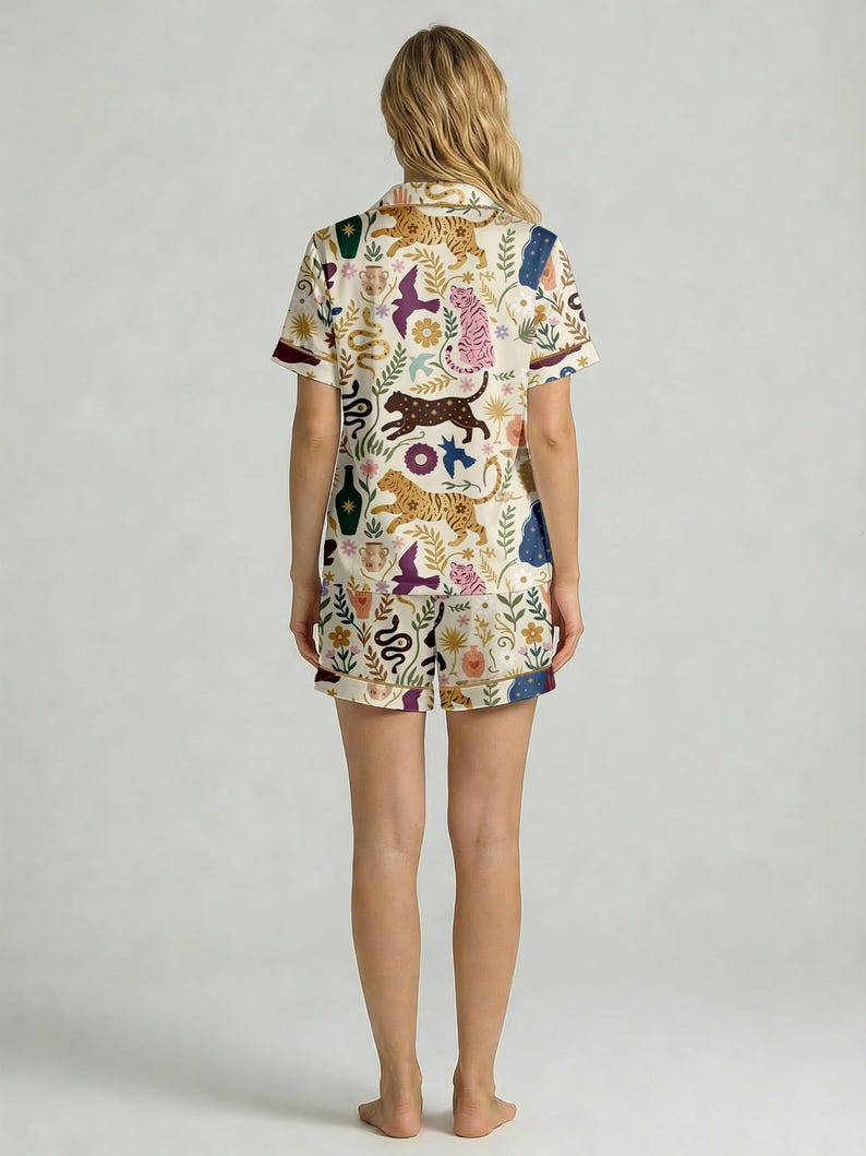 Pode incluir: Conjunto de pijama creme com um padr&atilde;o repetido de tigres, p&aacute;ssaros, cobras e elementos florais. A parte superior de manga curta e os cal&ccedil;&otilde;es t&ecirc;m uma borda contrastante. O design inclui cores castanho, rosa e azul.