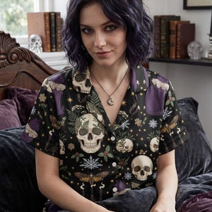Op de afbeelding: Zwart pyjamashirt met korte mouwen met een herhalend patroon van schedels, motten en botanische elementen. De top heeft een knoopsluiting aan de voorkant en een kraag. Het algemene thema is gotisch en grillig. Het model draagt een zilveren ketting.