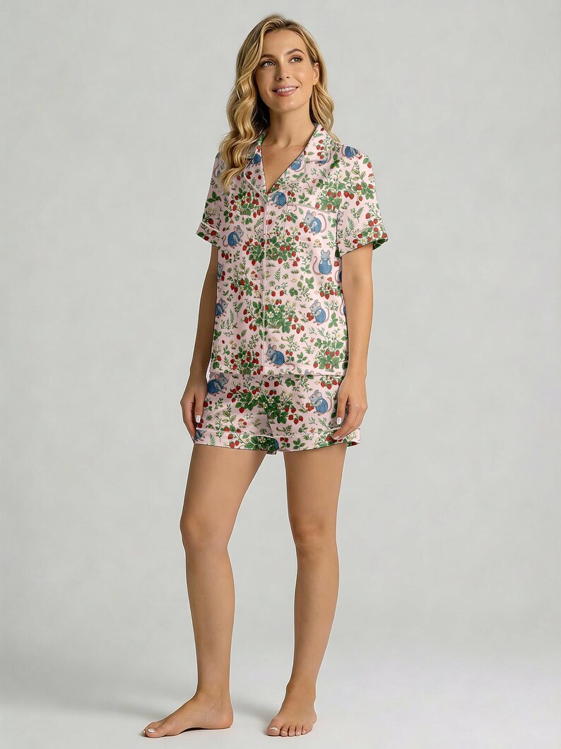 Puede incluir: Un conjunto de pijama rosa claro con un estampado de fresas y ratones. El conjunto incluye una camisa de manga corta con botones y pantalones cortos a juego. La tela tiene un ribete blanco y cuello en V.