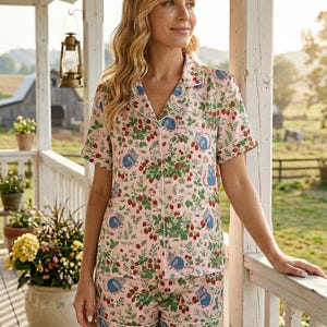 Puede incluir: Conjunto de pijama rosa con estampado de fresas y ratones. La parte superior de manga corta y los pantalones cortos tienen ribete blanco. El fondo muestra un porche, plantas y un granero.