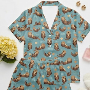 May include: A two-piece pajama set featuring a light blue background with a repeating pattern of brown otters, seashells, and wavy lines. The short-sleeved top has a collared neckline and button-down closure. The matching shorts have an elastic waistband.
