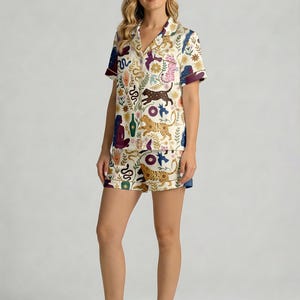 Pode incluir: Um conjunto de pijama de duas pe&ccedil;as creme. A parte de cima e os cal&ccedil;&otilde;es s&atilde;o adornados com um padr&atilde;o colorido de tigres, cobras e elementos florais. A parte de cima tem mangas curtas com acabamento roxo escuro, e os cal&ccedil;&otilde;es t&ecirc;m acabamento azul.