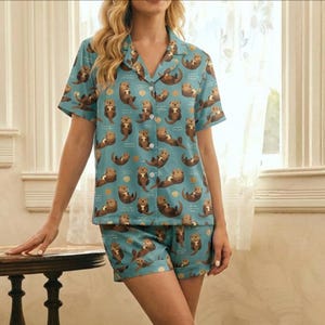 May include: A two-piece pajama set featuring a light blue background with a repeating pattern of brown otters and gold dots. The set includes a short-sleeved button-down shirt and matching shorts. The pajamas are made of a soft, lightweight fabric.