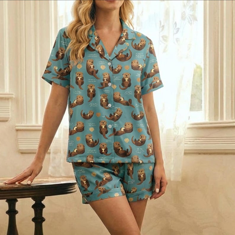 May include: A two-piece pajama set featuring a light blue background with a repeating pattern of brown otters and gold dots. The set includes a short-sleeved button-down shirt and matching shorts. The pajamas are made of a soft, lightweight fabric.
