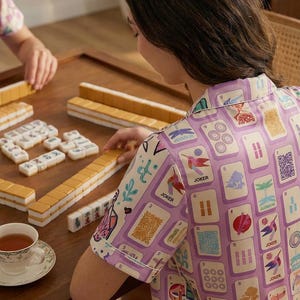Peut inclure: Une partie de Mahjong en cours. L'image montre une table en bois avec des tuiles et une tasse de thé. Une personne portant une chemise violette avec des illustrations de cartes joue au jeu.