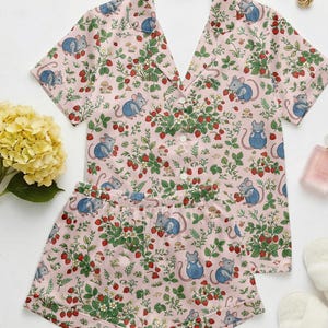 Puede incluir: Un conjunto de pijama rosa con un estampado de fresas, hojas y ratones de dibujos animados con monos azules. El conjunto incluye una camisa de manga corta con botones y pantalones cortos a juego. El pijama est&aacute; hecho de una tela suave y ligera.