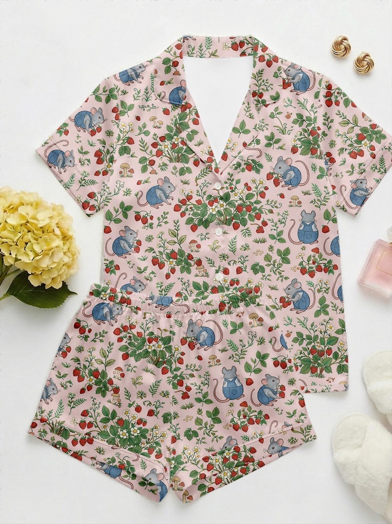 Puede incluir: Un conjunto de pijama rosa con un estampado de fresas, hojas y ratones de dibujos animados con monos azules. El conjunto incluye una camisa de manga corta con botones y pantalones cortos a juego. El pijama est&aacute; hecho de una tela suave y ligera.