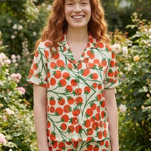 Puede incluir: Un conjunto de pijama color crema con un estampado repetido de tomates rojos y vides verdes. La parte superior de manga corta tiene un cuello con muescas y está abotonada. Los pantalones cortos tienen un estampado de tomate a juego.