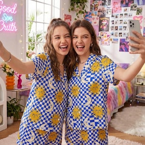 Op de afbeelding: Twee mensen dragen bijpassende blauw-witte geruite pyjamasets met gele zon- en maangraphics. Ze lachen en maken een selfie met een telefoon. De achtergrond omvat een neonbord en een kleurrijk sprei.
