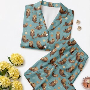 May include: A teal pajama set featuring a repeating pattern of brown otters, seashells, and wavy lines. The top has a collared neckline and button closure. The pajama bottoms have an elastic waistband. The set is made of a soft, comfortable material.