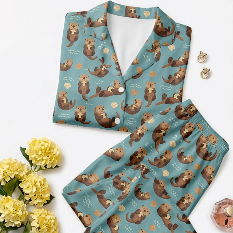 May include: A teal pajama set featuring a repeating pattern of brown otters, seashells, and wavy lines. The top has a collared neckline and button closure. The pajama bottoms have an elastic waistband. The set is made of a soft, comfortable material.