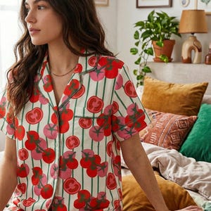 Può includere: Un completo pigiama a maniche corte con un motivo di pomodori rossi e rosa su sfondo bianco a righe verdi. Il completo comprende una camicia abbottonata e pantaloncini, con bordi verdi.