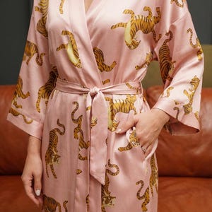 May include: A light pink satin robe featuring a repeating pattern of orange and black tigers. The robe has a tie waist, long sleeves, and pockets. The robe is a luxurious and comfortable garment.