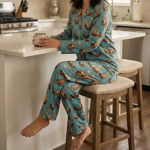 May include: Teal pajama set featuring a repeating pattern of brown otters. The long-sleeved top has a button-down front and a notched collar. The matching pants have an elastic waistband. The person is sitting on a wooden stool.