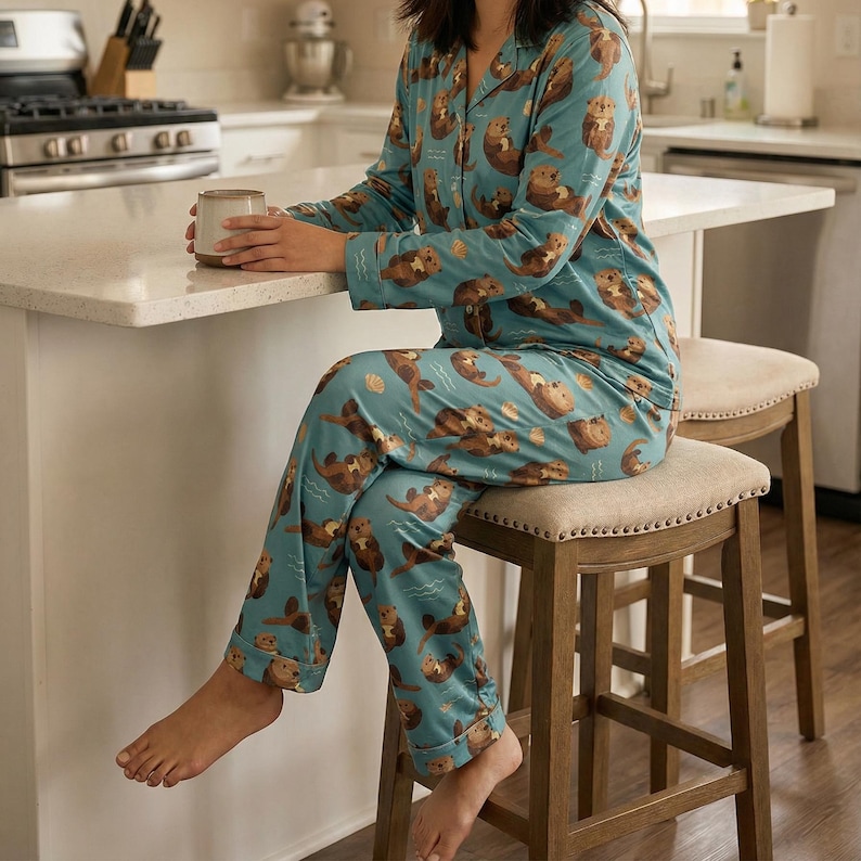 May include: Teal pajama set featuring a repeating pattern of brown otters. The long-sleeved top has a button-down front and a notched collar. The matching pants have an elastic waistband. The person is sitting on a wooden stool.