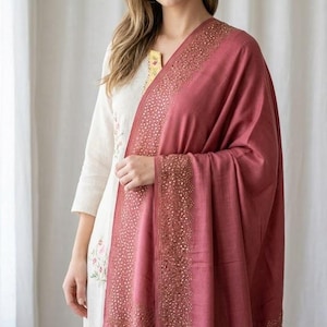 May include: A woman wearing a cream-colored embroidered tunic and a draped, dusty rose shawl. The shawl features a detailed, gold-toned embellished border. The outfit is set against a soft, neutral background.