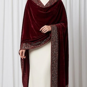 Puede incluir: Un chal de terciopelo color burdeos intenso con un borde ancho adornado con intrincados abalorios y lentejuelas en tonos dorados. La prenda se coloca sobre un vestido largo de color crema, destacando una textura rica y una caída pesada.
