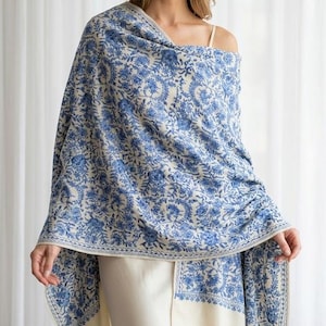 Può includere: Uno scialle rettangolare caratterizzato da una base color crema con un intricato motivo floreale blu. Il design include dettagliati motivi botanici e un bordo decorativo lungo i lati. Il tessuto si drappeggia sulle spalle e sulla parte superiore del corpo.