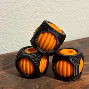 May include: Three 3D-printed geometric objects. Each object has a black, faceted outer shell with orange, ribbed spheres visible through the openings. The objects are stacked on a dark wooden surface, with a neutral background.