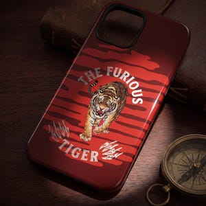 Könnte beinhalten: Eine weinrote Handyhülle mit einer Tigerillustration und dem Text "THE FURIOUS TIGER". Die Hülle hat einen rot gestreiften Hintergrund und Krallen. Auf der Unterseite der Hülle steht der Text "GHOST ENGRAVING" und "VISIBLE IN DIRECT LIGHT | INVISIBLE IN SHADOW".