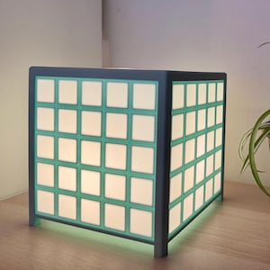 May include: A cube-shaped lamp with a grid pattern of white squares and teal borders. The lamp is illuminated, casting a soft glow. A potted plant with long green and white leaves sits nearby, adding a touch of nature to the scene.