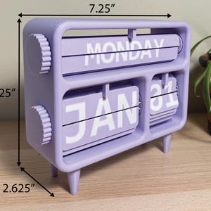 Pu&ograve; includere: Un calendario perpetuo da scrivania color lavanda. Mostra "LUNED&Igrave;" e "31 GEN" in bianco. Il calendario &egrave; largo 18,4 cm, alto 15,9 cm e profondo 6,7 cm. Ha due manopole circolari e quattro piedini.