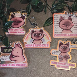 Può includere: Una collezione di portachiavi a tema gatto con varie frasi e illustrazioni. Ogni portachiavi presenta un gatto dei cartoni animati con diverse espressioni e testo come "Executive Dysfunction", "Hyperfocus" e "Time Blindness".