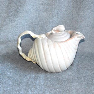 May include: A cream-colored teapot with a seashell design. The teapot has a curved handle and a spout, and the lid is topped with a small, decorative element. The teapot is made of ceramic or porcelain.