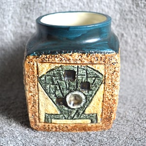 May include: A square ceramic vase with a textured, tan exterior and a teal blue rim. The front features a geometric design in green and tan, with a clear, round embellishment in the center. The interior is white.