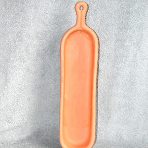 May include: An orange-colored, rectangular serving tray with a handle and a hole for hanging. The tray is made of a solid material and has a smooth surface. The handle is rounded and has a small hole at the top. The tray is designed for serving food or displaying items.
