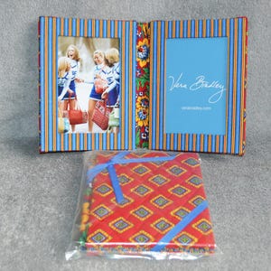 May include: A Vera Bradley photo frame with a blue and yellow striped pattern, open to display a photo and a blue background with the Vera Bradley logo. A red patterned item is in the foreground.