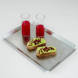 Può includere: Un vassoio rettangolare in acciaio inossidabile con bordi ripiegati. Contiene due bicchieri alti e trasparenti pieni di liquido rosso e due fette di toast con avocado e semi di melograno. Il vassoio è su una superficie bianca.