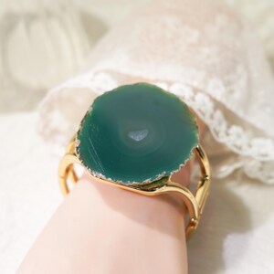 Puede incluir: Un brazalete dorado con una gran rodaja de ágata verde pulida y circular como pieza central. La piedra muestra bandas concéntricas naturales y una superficie lisa, montada de forma segura sobre una banda de metal pulido con un diseño abierto.
