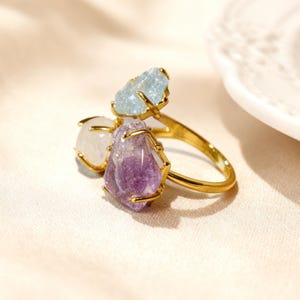 Op de afbeelding: Een goudkleurige ring met drie ruwe, ongeslepen edelstenen die op hun plaats worden gehouden door metalen pootjes. De stenen omvatten een lichtblauw kristal, een doorschijnend wit kristal en een paarse amethist op een dunne, gepolijste ring.