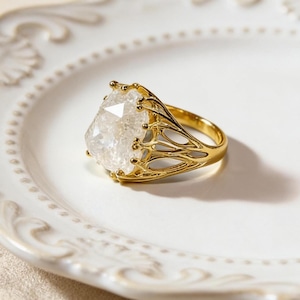 May include: A gold ring featuring a large, clear, faceted gemstone. The ring has an elaborate, openwork design around the stone and a smooth, polished band. The ring is displayed on a white, decorative plate.