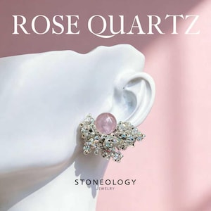 Puede incluir: Un pendiente de cuarzo rosa con una gran gema rosa en el centro, rodeada por un grupo de piedras más pequeñas y transparentes. El pendiente se muestra en una cabeza de maniquí blanca sobre un fondo rosa. Los textos "ROSE QUARTZ" y "STONEOLOGY JEWELRY" son visibles.