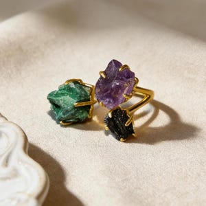Peut inclure: Trois bagues dorées, chacune ornée d'une pierre brute différente. Une bague présente une pierre verte, une autre une pierre violette et la troisième une pierre noire. Les pierres précieuses de forme irrégulière ajoutent une esthétique naturelle et organique aux bijoux.