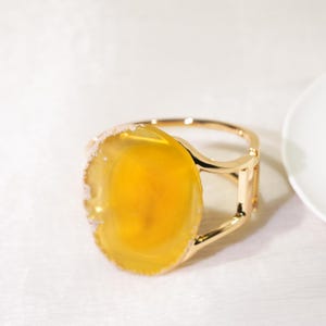 May include: A gold-toned metal cuff bracelet featuring a large, circular, translucent yellow stone centerpiece. The stone has a smooth, polished surface with a natural, textured, and slightly irregular white edge, set within a polished gold frame.