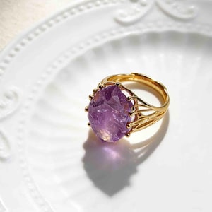 May include: A gold ring with a large, raw amethyst gemstone. The amethyst is a deep purple colour with a faceted surface. The ring has an ornate, openwork design around the gemstone.