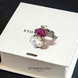May include: A silver ring with a large, rough-cut, magenta gemstone and a white, baroque pearl. The ring is displayed on a white box with the text "STONEOLOGY JEWELRY".