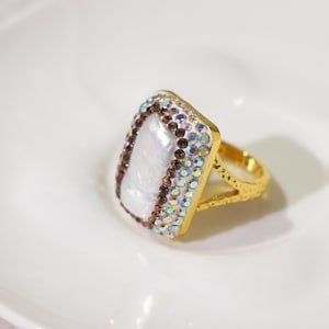 May include: A gold-toned ring featuring a rectangular central white iridescent stone. The stone is bordered by a row of small dark brown crystals and an outer row of shimmering multi-colored iridescent crystals on a textured gold band.