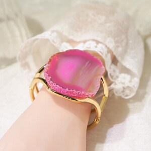 May include: A gold-toned cuff bracelet featuring a large, polished, vibrant pink agate slice as the centrepiece. The natural stone has a translucent, banded appearance with a textured, crystalline edge, securely mounted on a smooth, curved metal band.
