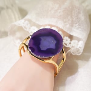 Puede incluir: Un brazalete tipo puño de tono dorado que presenta una gran rodaja de ágata circular de color púrpura intenso como punto focal central. La banda de metal tiene un acabado liso y pulido con un diseño abierto que sujeta la piedra en el centro.