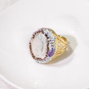 May include: A gold-toned ring featuring a large, iridescent, oval-shaped central stone surrounded by a border of small, sparkling, multi-colored crystals and brown stones. The band has an open, intricate, wire-like design in a metallic gold finish.