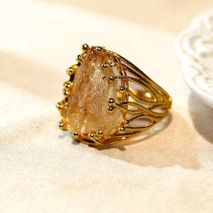 May include: A gold-colored ring featuring a large, irregularly shaped, light-brown gemstone. The ring has a decorative, openwork design with small gold beads around the gemstone and along the band. The ring is set on a cream-colored surface.