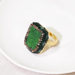 May include: A gold-toned ring featuring a large, rectangular, textured green druzy stone centrepiece. The stone is framed by a border of small, dark green crystals and a thin outer edge of light brown, shimmering, faceted stones on a gold band.